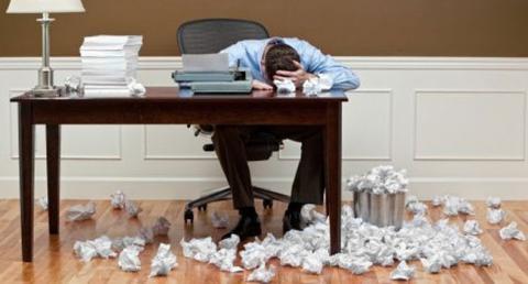 Man with his head on a desk, surrounded by crumpled balls of paper