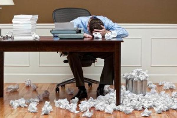 Man with his head on a desk, surrounded by crumpled balls of paper